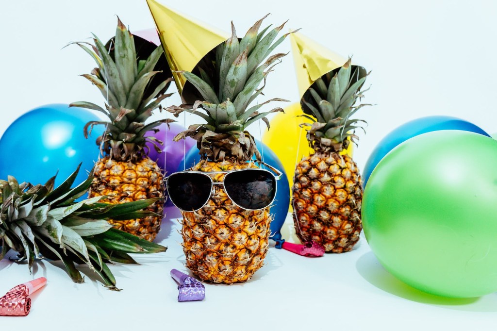 A pineapple with sunglasses, with festive hats, balloons, and streamers. 